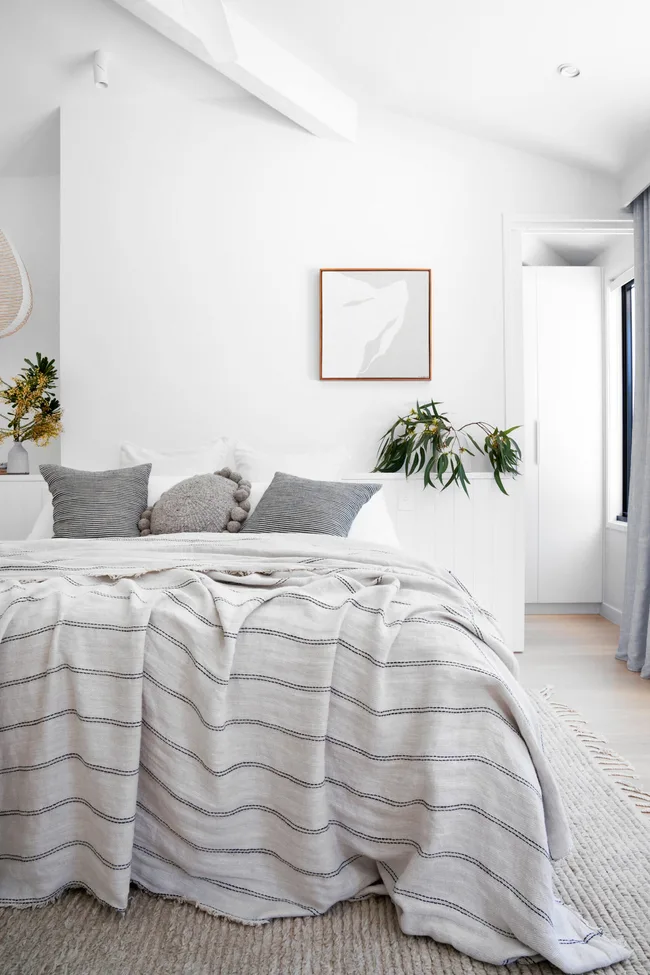 Cozy bedroom with a striped blanket, gray and white pillows, abstract wall art, and greenery on a light-filled white wall.