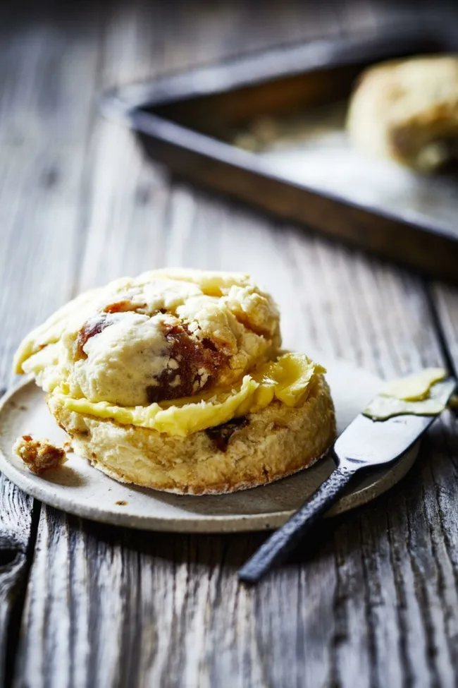 A freshly baked scone with butter on a wooden surface next to a knife.