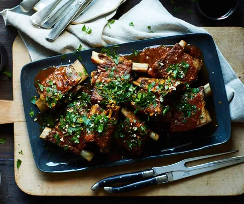 Braised short ribs on a dark platter, garnished with fresh herbs, set on a wooden board with cutlery nearby.
