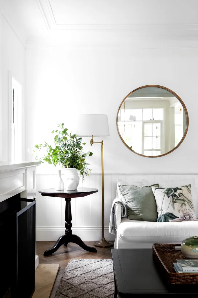 Cozy living room with round mirror, white sofa, green pillows, small table with plants, and a floor lamp.