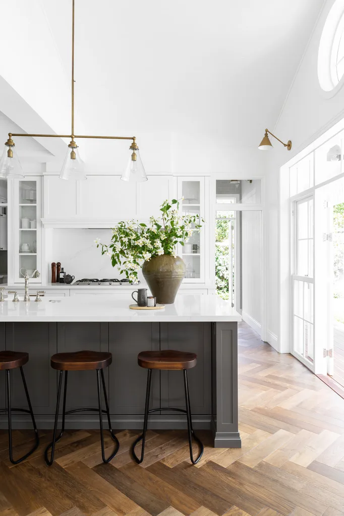 Bright kitchen with white cabinets, wooden stools at island, pendant lights, and a vase with greenery on the counter.