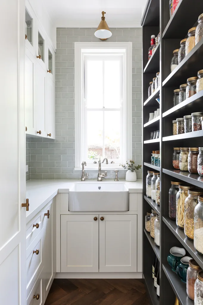 Pantry with white cabinets, farmhouse sink, gray tile backsplash, and shelves filled with jars and kitchen items.