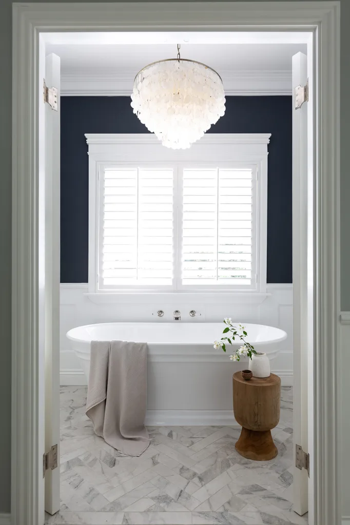Elegant bathroom with a freestanding tub, chandelier, wood stool, and flowers, framed by open white doors.