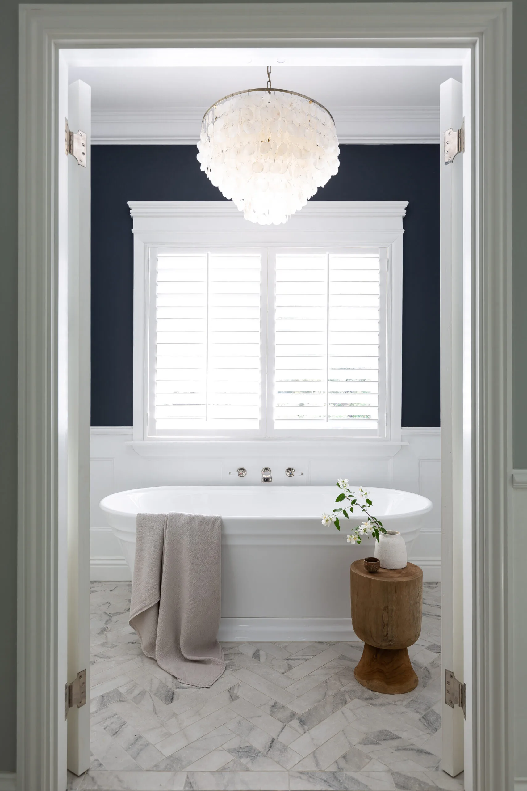 Elegant bathroom with a freestanding tub, chandelier, wood stool, and flowers, framed by open white doors.