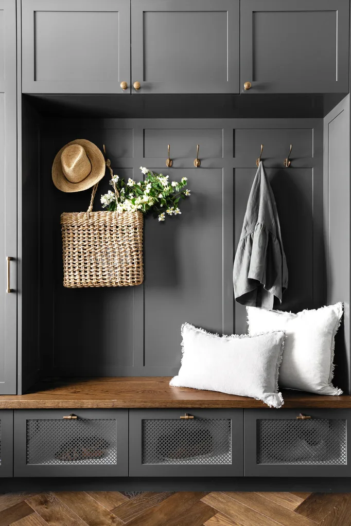 Grey mudroom entryway with brass clothes hooks and timber bench seating, with white cushions, straw bag and hat.