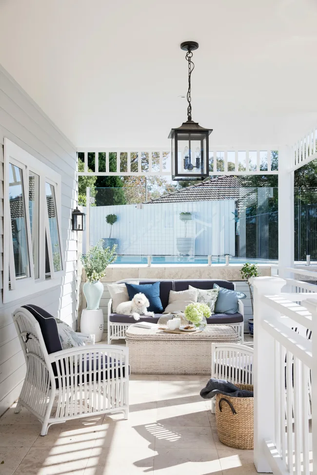 Covered patio with wicker furniture, cushions, a white fluffy dog, and a lantern above. Pool visible in the background.