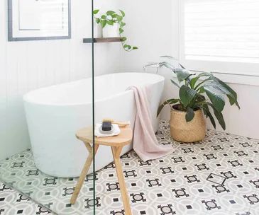 Modern bathroom with freestanding tub, plant, and patterned tiles; wooden stool with soap and brush.
