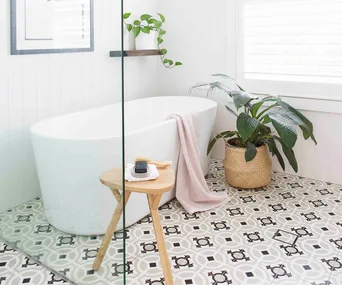Modern bathroom with freestanding tub, plant, and patterned tiles; wooden stool with soap and brush.