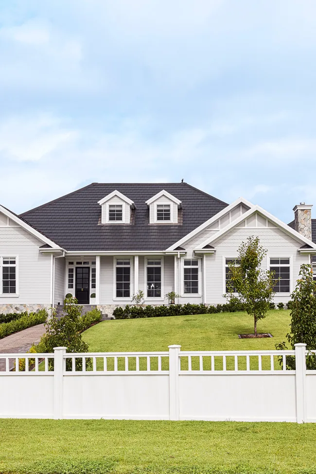 A light-colored house with a dark roof, front lawn, and white fence under a clear blue sky.