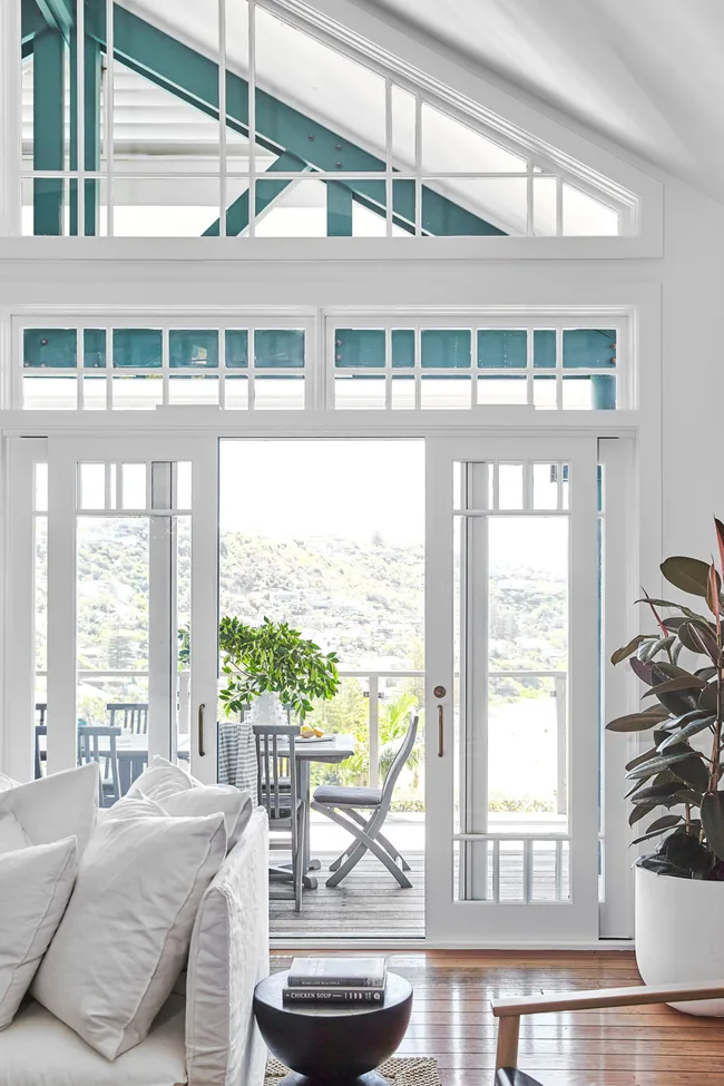 Bright living room with white furniture, large windows, wooden floors, and a view of an outdoor patio with greenery.
