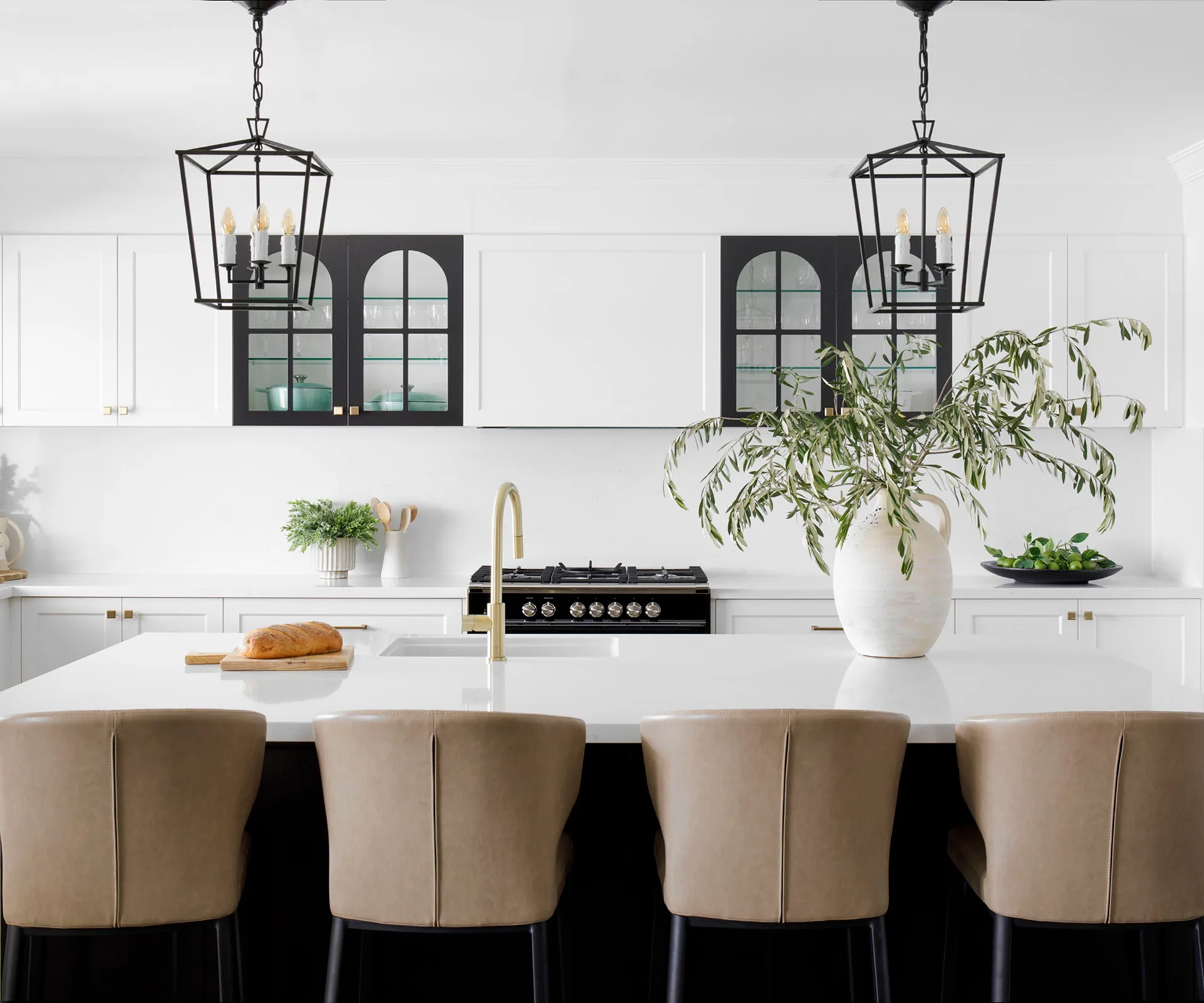 Modern kitchen with white cabinets, black stove, large island with beige chairs, two pendant lights, and a plant on the counter.