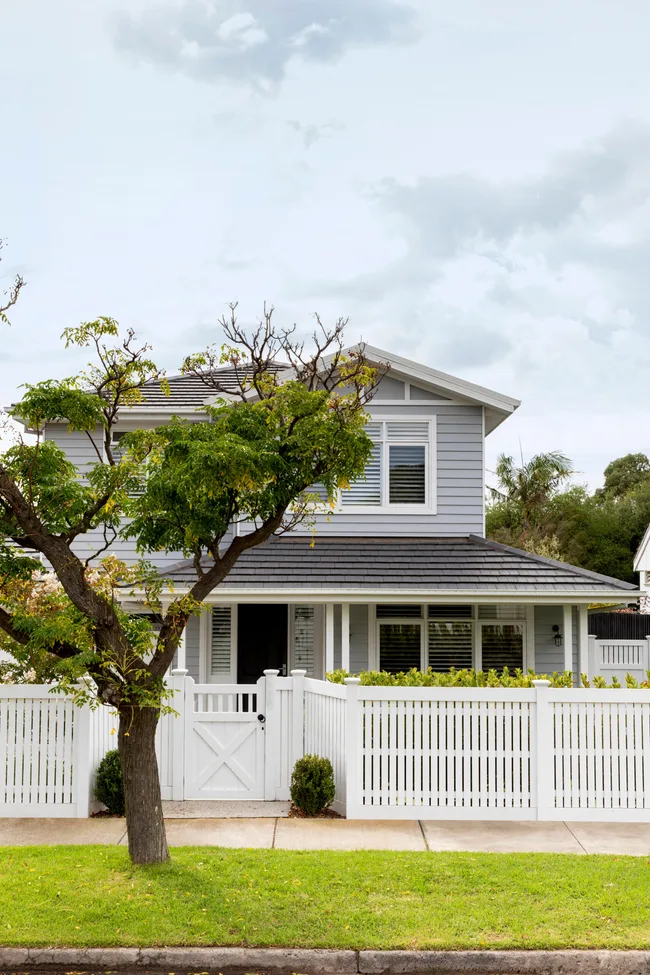 Two-story gray house with white fence, surrounded by green trees and a well-kept lawn, under a cloudy sky.