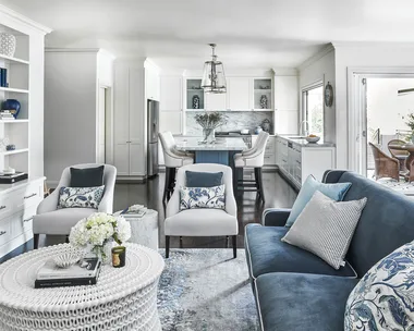 Elegant living room and kitchen with blue and white decor, featuring a sofa, armchairs, and a white wicker coffee table.