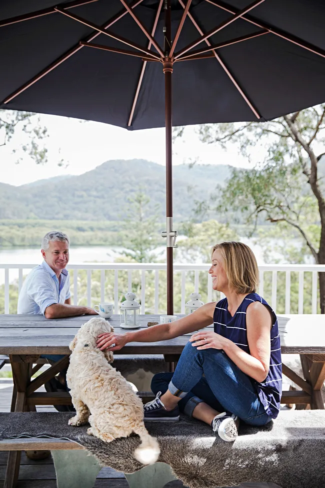 Couple sitting under outdoor umbrella with a dog, trees and mountains in background.