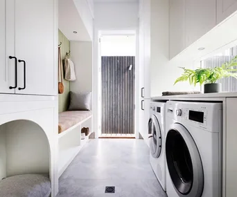 Laundry room with white cabinets, washer and dryer. Bench with green accents and a door leading outside.