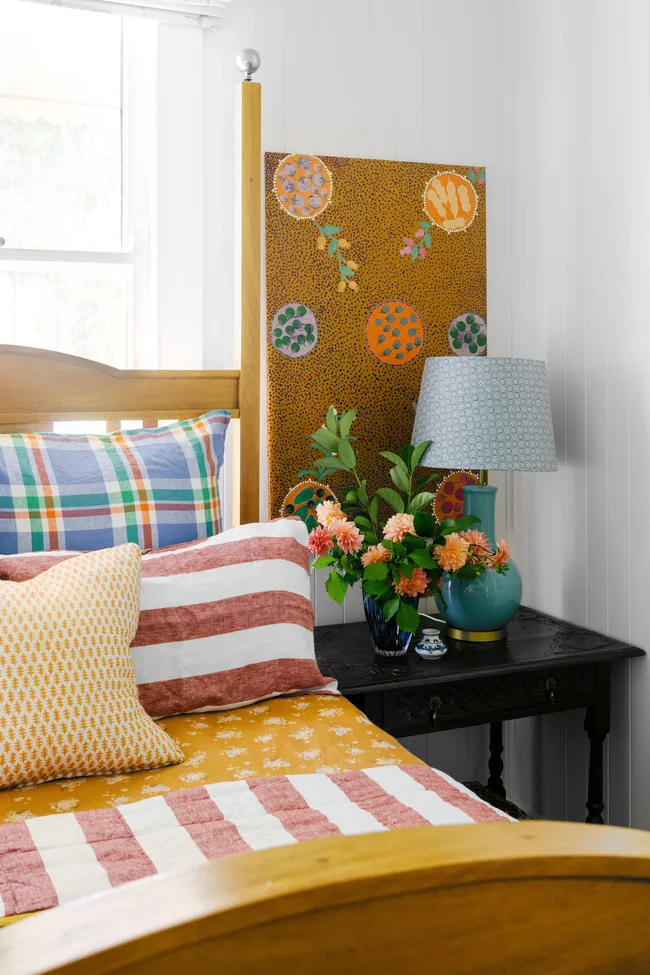 Cozy bedroom with colorful pillows, a floral quilt, abstract art, and a bedside table with a lamp and flowers.