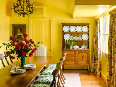 Yellow dining room with wooden table, floral chairs, chandelier, and hutch of plates. Vase with red flowers on table.