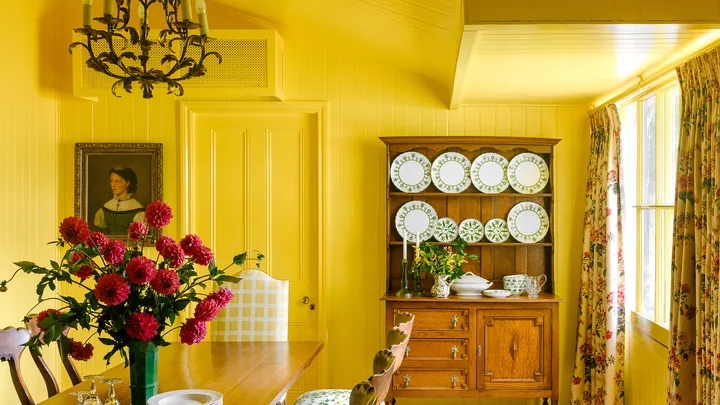 Yellow dining room with wooden table, floral chairs, chandelier, and hutch of plates. Vase with red flowers on table.