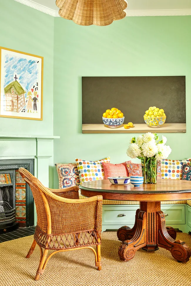 Dining area with a wicker chair, round wooden table, colorful pillows, vase of flowers, and fruit paintings on mint walls.