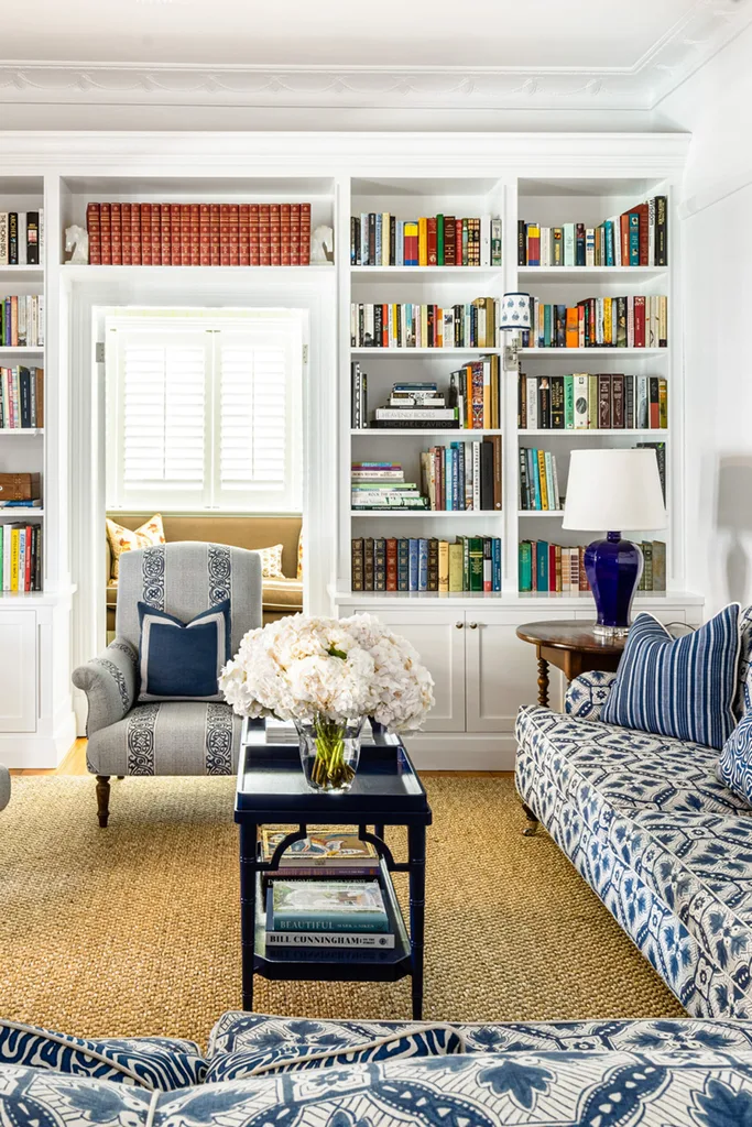 Living room with bookshelves, blue-patterned sofa, armchair, and table with white flowers and lamp.