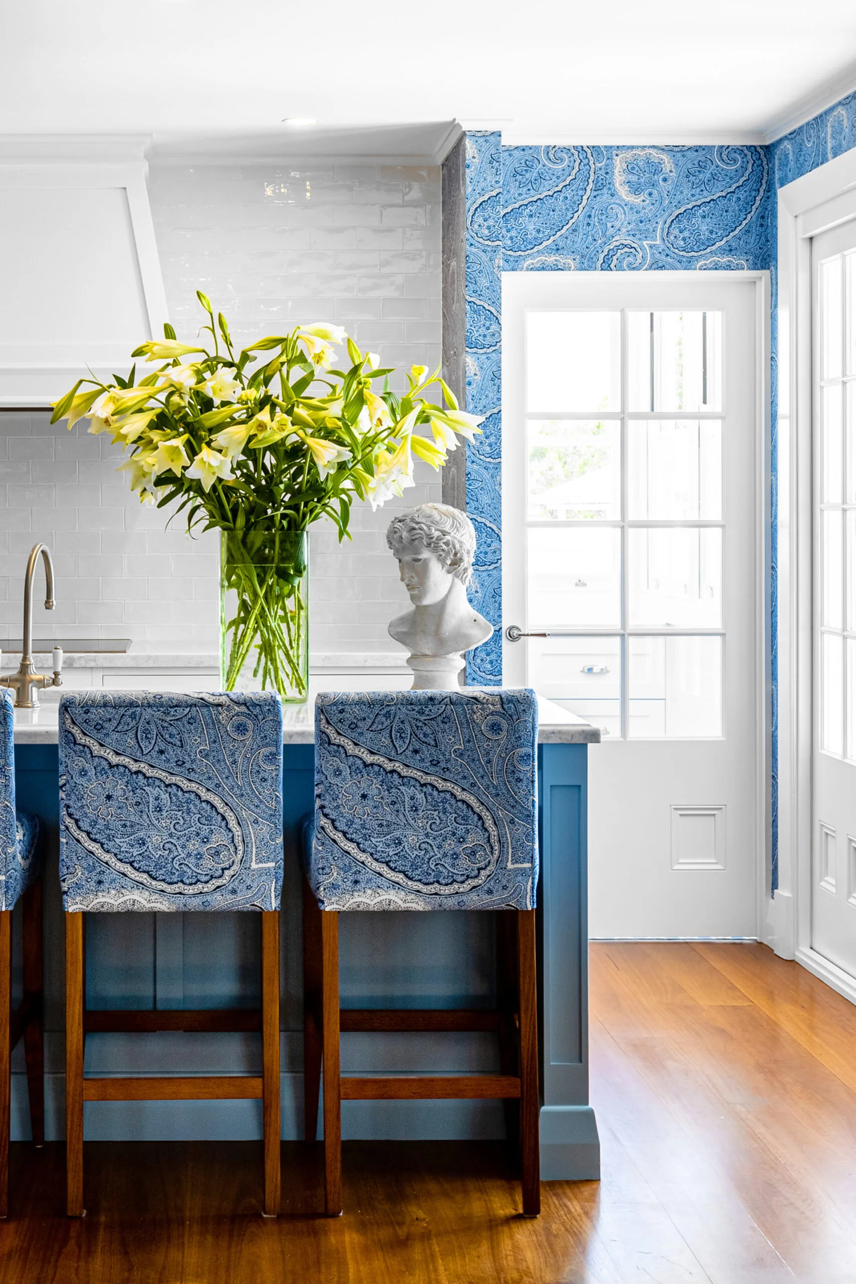 Paisely blue wallaper and matching upholstered bar stools in a white and blue kitchen