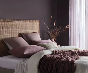 Cozy bedroom with rattan headboard, soft pillows, mauve knitted blanket, and light filtering through sheer curtains.