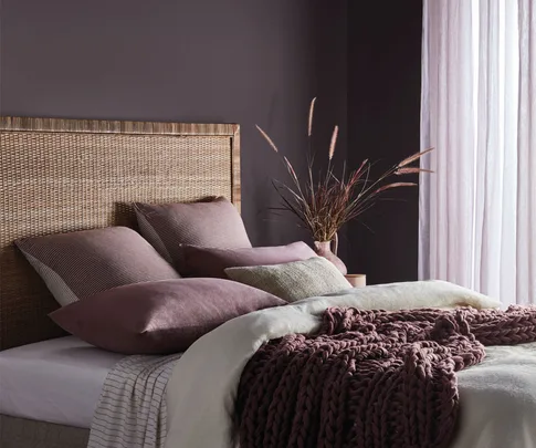 Cozy bedroom with rattan headboard, soft pillows, mauve knitted blanket, and light filtering through sheer curtains.
