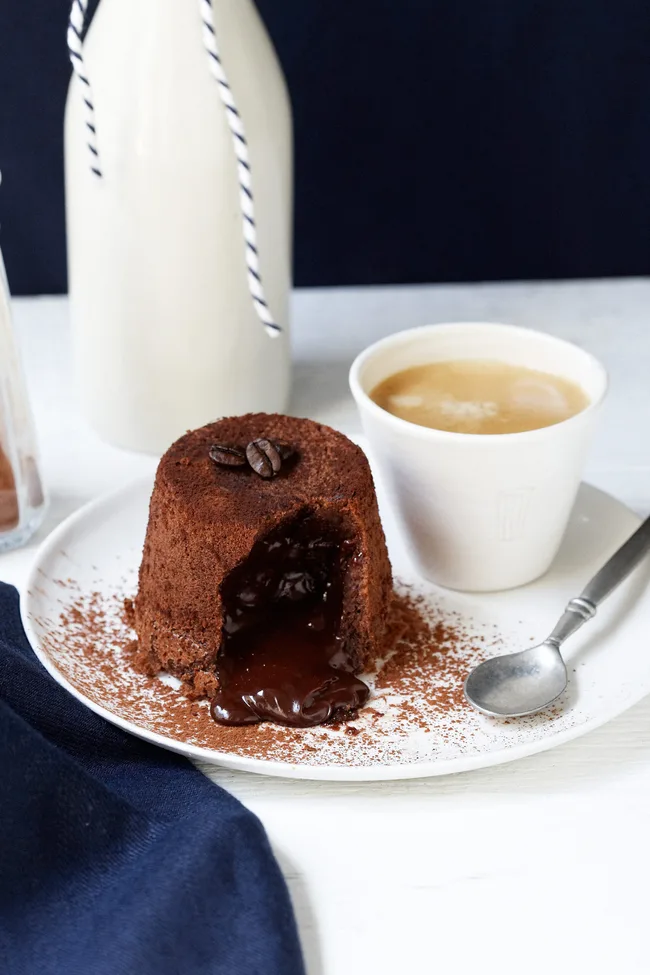 Chocolate lava cake with melted centre on a plate, topped with coffee beans, next to a cup of coffee and a spoon.