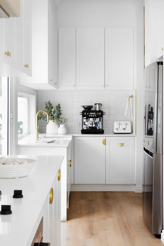 A modern, white kitchen with wooden floors, featuring a coffee machine, toaster, gold fixtures, and minimal decor.