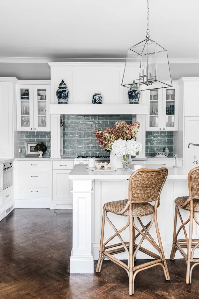 Elegant white modern hamptons kitchen with green tile backsplash, wicker bar stools, and glass pendant light over island.