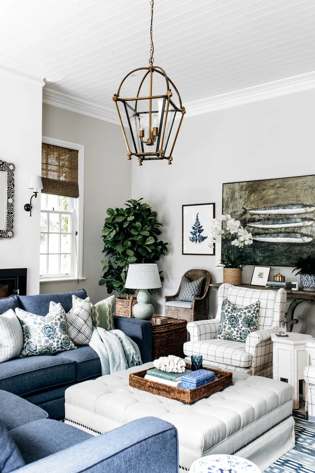 Living room with blue sofas, patterned pillows, large ottoman, indoor plant, decorative art, and pendant light fixture.