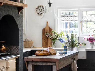 Rustic kitchen with a wooden island, bread, flowers, vintage stove, and a hanging glass light fixture.