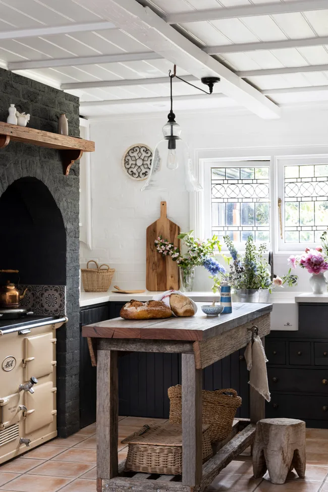 Rustic kitchen with a wooden island, bread, flowers, vintage stove, and a hanging glass light fixture.