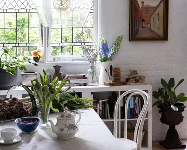 Cozy dining area with glass light fixture, plants, books, and a tea set on the table by a window with stained glass details.