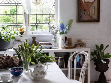 Cozy dining area with glass light fixture, plants, books, and a tea set on the table by a window with stained glass details.