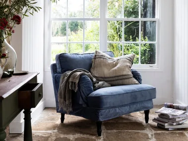 Cozy room with a blue armchair, cushions, and a blanket by a large window; plants and magazines nearby.
