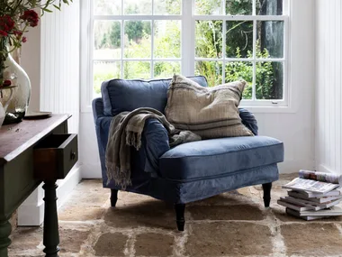 Cozy room with a blue armchair, cushions, and a blanket by a large window; plants and magazines nearby.