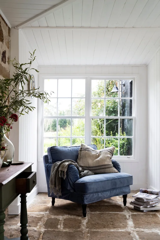 Cozy room with a blue armchair, cushions, and a blanket by a large window; plants and magazines nearby.