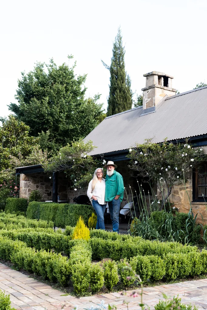 Couple standing in front of a rustic stone house surrounded by lush green garden and trees.