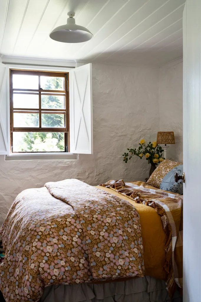 Cozy bedroom with a floral quilt, a window with shutters open, and a bedside lamp with yellow flowers in a vase.