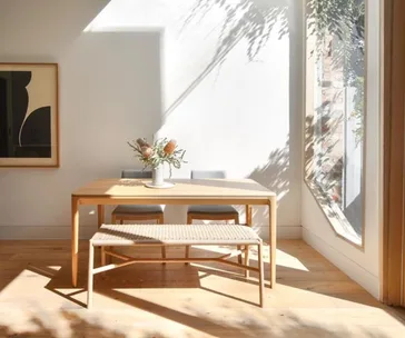Sunlit dining area with a wooden table, two chairs, and a bench. A vase with flowers sits on the table. Art on the wall.