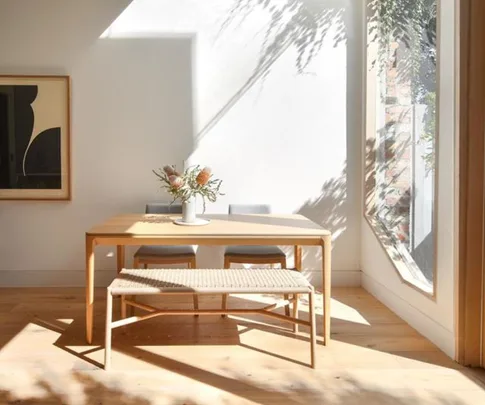 Sunlit dining area with a wooden table, two chairs, and a bench. A vase with flowers sits on the table. Art on the wall.