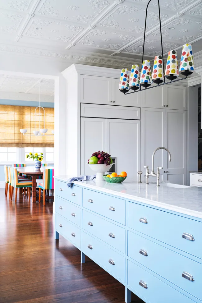 Kitchen with blue island, colorful pendant lights, fruit bowl, and white cabinets, leading to a dining area with striped chairs.