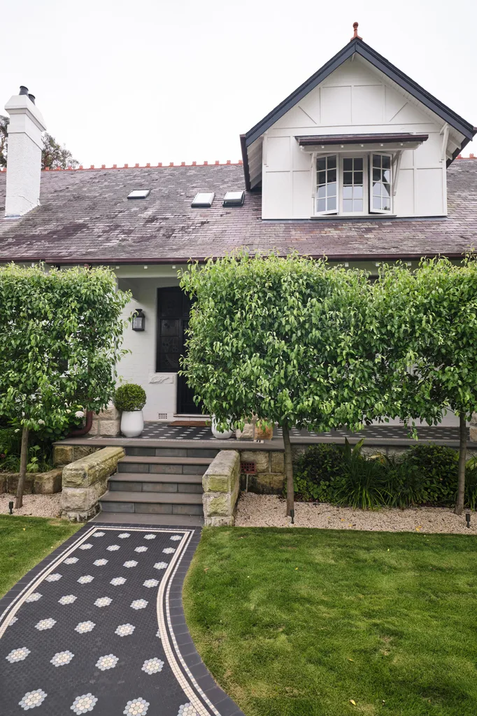 Charming house with slate roof, dormer window, lush trees, and a decorative tiled pathway on a well-manicured lawn.