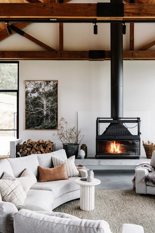 Cozy living room with a light gray curved sofa, pillows, a white round table, and a lit fireplace. Logs and artwork nearby.