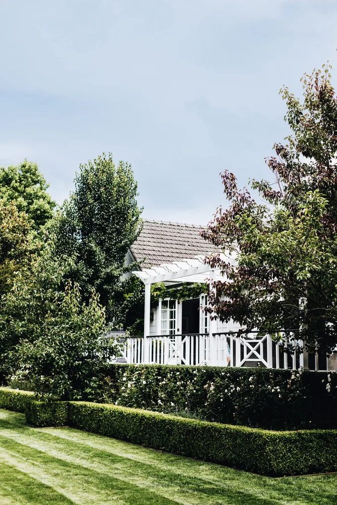 Charming white cottage with lush green garden, porch, and hedges under a clear blue sky.