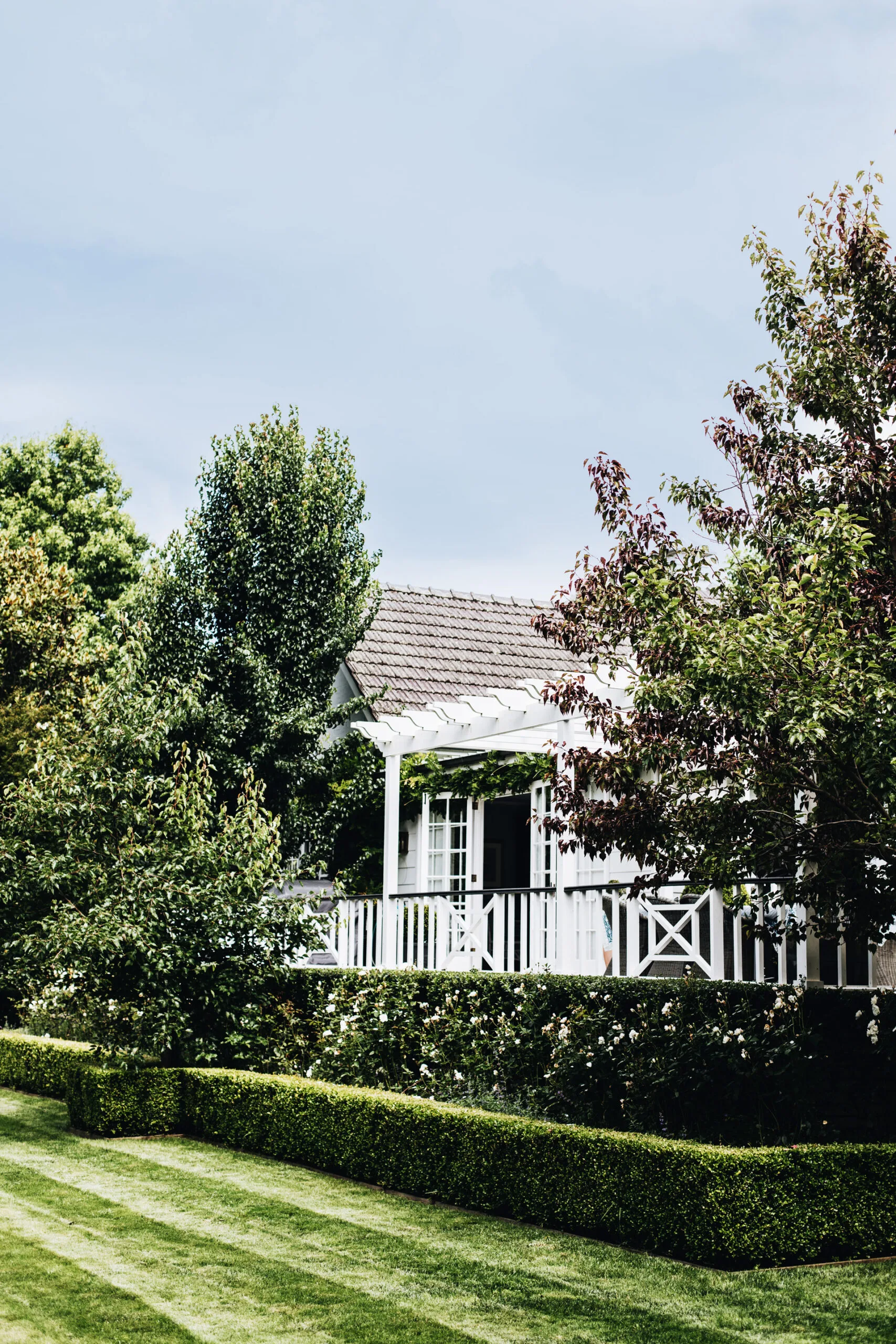 Charming white cottage with lush green garden, porch, and hedges under a clear blue sky.