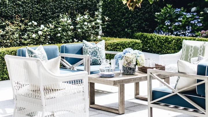 Outdoor patio with white and blue furniture, surrounded by greenery and flowers, under a tree canopy.