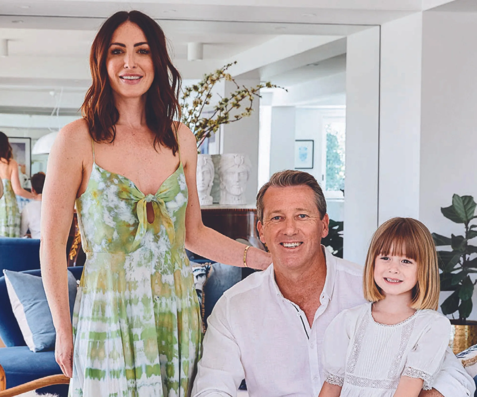 A woman in a green dress, a man in a white shirt, and a young girl in a white dress smile in a bright, modern room.