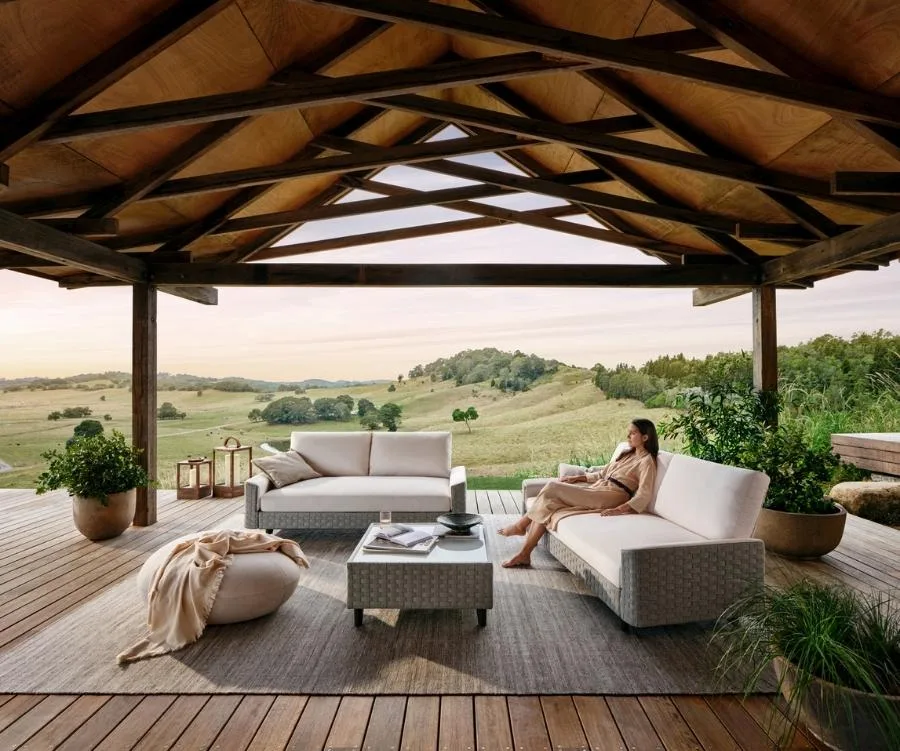 Outdoor patio with sofa, table, and a person seated, overlooking a scenic landscape of rolling hills and greenery.